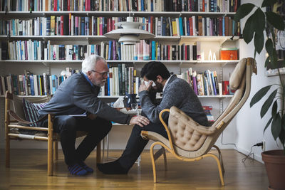 Senior therapist counseling young patient while sitting by bookshelf during therapy session