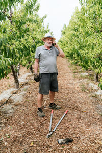 Full length senior man in casual clothes and hat standing near shears on path amidst fruit trees and talking on cellphone during work on farm on summer day