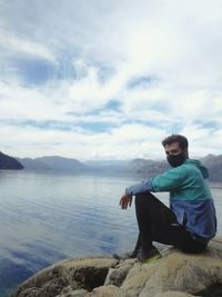 Man sitting on rock by lake against sky