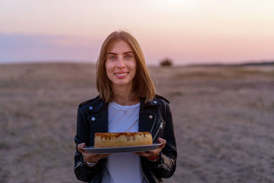 Portrait of smiling young woman holding cake against sky during sunset