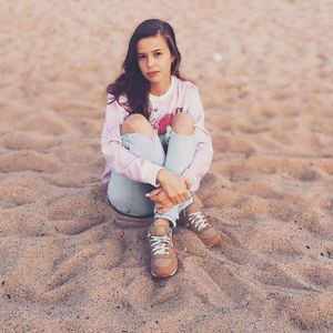 Portrait of young woman hugging knees while sitting on sand at beach