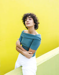 Self assured curly haired female folding hands while looking at camera with serious glance against bright yellow background