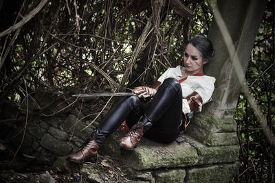 Full length of woman wearing costume with sword sitting at forest