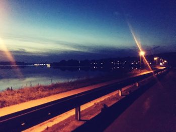 Illuminated street by river against sky at night