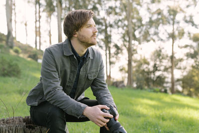 Portrait of young man sitting on tree stump outside holding camera