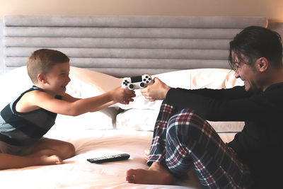 Father and son sitting on a bed and struggling over video game controller.