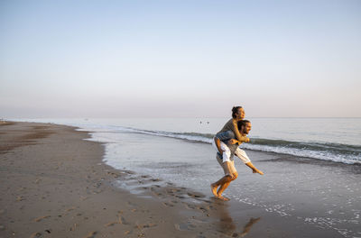 Cheerful man giving piggyback to girlfriend on shore at beach against clear sky during sunset