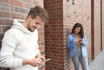 Full length of young man holding smart phone while standing against brick wall