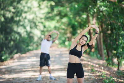 Man and woman stretching arms while exercising on road amidst trees