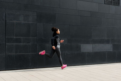 Side view of woman running on footpath by black wall