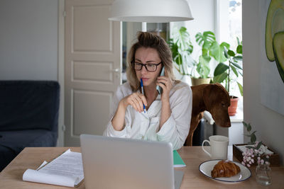 Employee woman in pajamas sitting with dog in living room taking on cellphone working at laptop