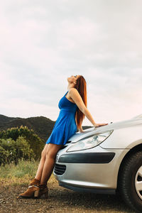 Side view of young woman standing on car against sky