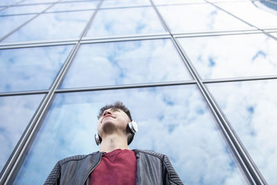 Young man listening music through headphones against glass wall