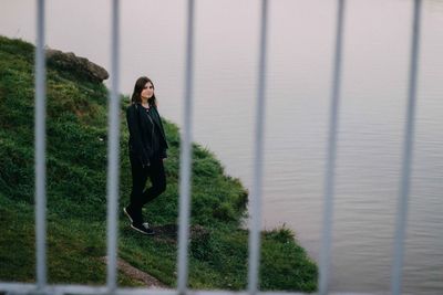 Full length of young woman standing by lake seen through railing