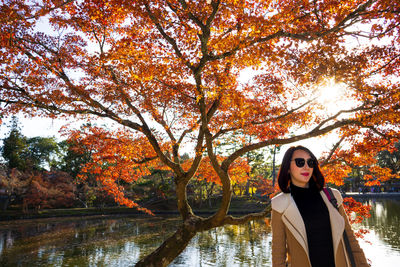 Portrait of young woman standing by tree against lake during autumn