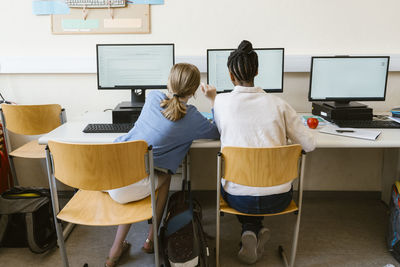 Rear view of girl assisting female friend using computer while sitting on chair in classroom at school