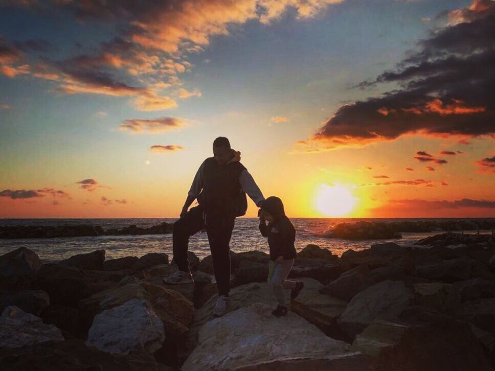 FRIENDS STANDING ON ROCK AT BEACH DURING SUNSET