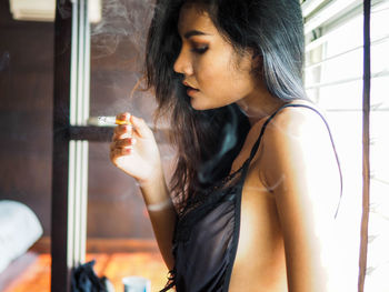 Side view of sensuous young woman smoking cigarette against window