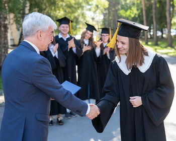 The teacher shakes hands with the student and presents the diploma outdoors. a group of university