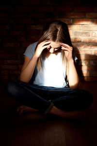 Young woman with headache while sitting at home by brick wall