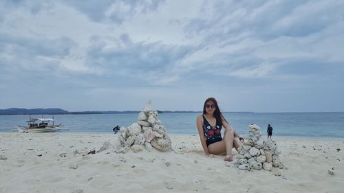 Portrait of smiling woman sitting by stacked rocks at beach against sky