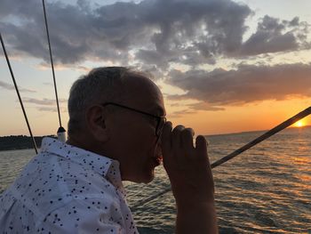 Side view of man having drink by sea against cloudy sky during sunset