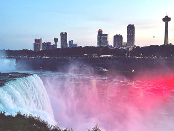 Niagara falls at dusk with red lights shining into the mist under a blue sky
