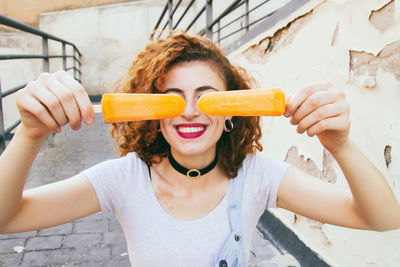 Close-up of woman smiling while holding orange popsicles on footpath