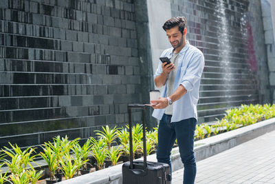 Young man using mobile phone while standing against wall