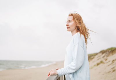 Portrait of young red haired woman in light blue sweater on sand beach by sea in storm
