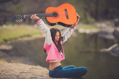 Frustrated young woman holding guitar while sitting at lakeshore