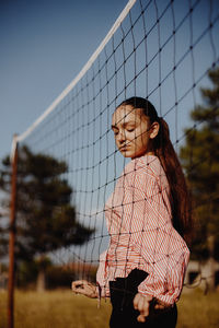 Side view of young woman standing by volleyball net against blue sky