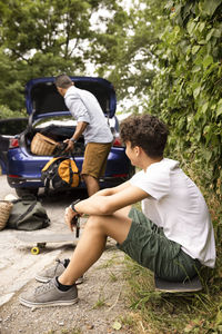 Side view of teenage boy sitting on skateboard looking at father loading luggage in car trunk