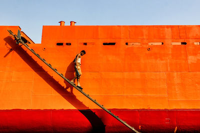 Side view of man walking on ladder by wall against sky