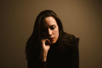 Unhappy pensive young female in black wear leaning on hand and thinking about personal problems while sitting against brown wall