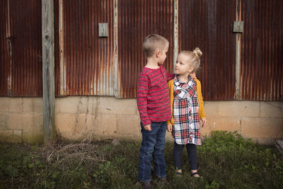 Siblings looking each other face to face while standing against abandoned building on field