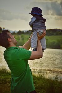 Father lifting son at riverbank against cloudy sky