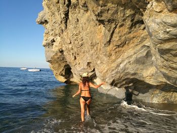 Rear view of sensuous woman walking on sea by rock formation at beach