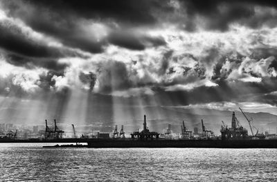 Cranes at harbor by sea against cloudy sky