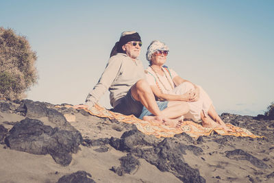 Happy senior couple sitting at beach against sky