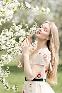 A young blonde in a long white dress poses near a cherry blossom in the garden, a spring landscape.
