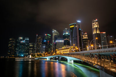 Illuminated bridge over river by buildings against sky at night