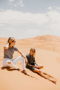 Friends sitting on sand dune in desert against sky