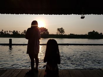Rear view of silhouette woman standing by lake against sky during sunset