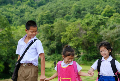 Siblings in school uniforms holding hands while standing against trees