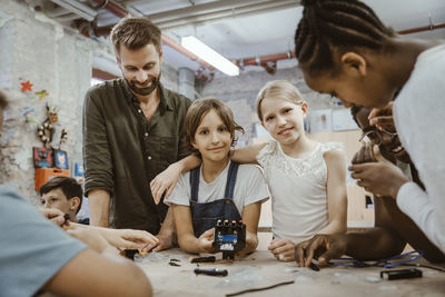 Portrait of smiling girl with arm around female friend amidst teacher and pupils at technology workshop in school