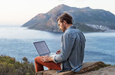 Side view of man using laptop on hill against mountain and sea