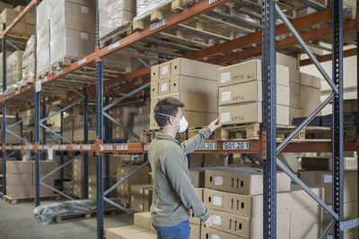 Man wearing mask holding boxes on shelf in warehouse