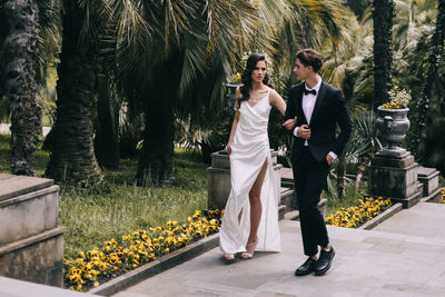 Happy lovers the bride and groom in wedding outfits walk among plants and palm trees in the old park