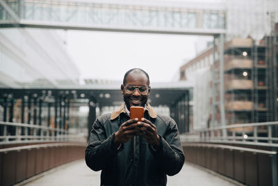 Smiling businessman using smart phone while standing on footbridge against sky in city